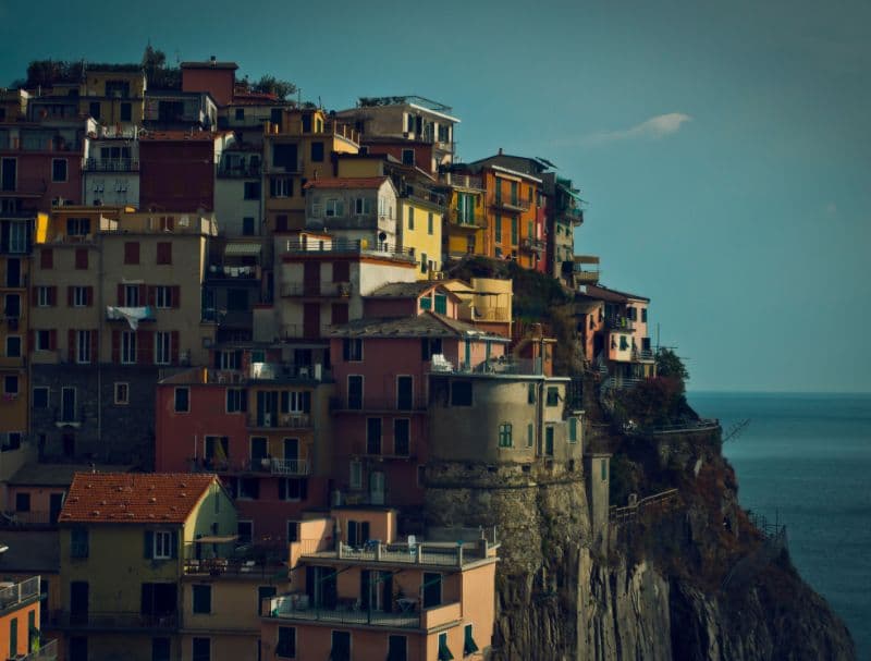 A picturesque view of the colorful hillside village in Cinque Terre, Italy. Multicolored buildings are built into the steep cliff face, overlooking the deep blue Ligurian Sea under a slightly hazy sky.