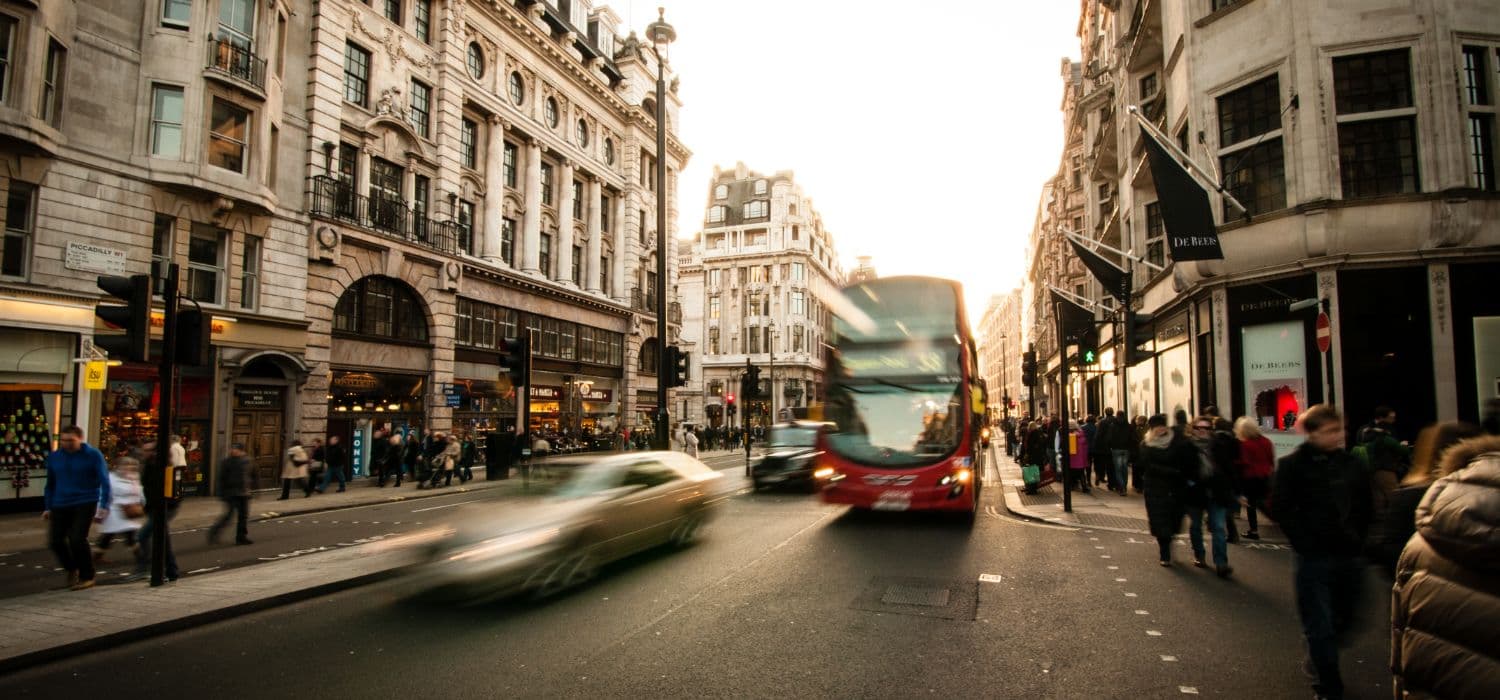 Street level view of a bustling London thoroughfare flanked by traditional multi-story buildings. A red double-decker bus is prominent in the center, moving towards the viewer, alongside other traffic including a motion-blurred car in the foreground. Pedestrians populate the sidewalks under a bright sky.