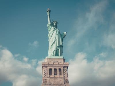 The Statue of Liberty is shown from the chest up, standing on its brown stone pedestal. The iconic copper-green statue holds a golden torch aloft in its right hand and a tablet in its left. Behind the statue, the sky is blue with scattered white clouds.